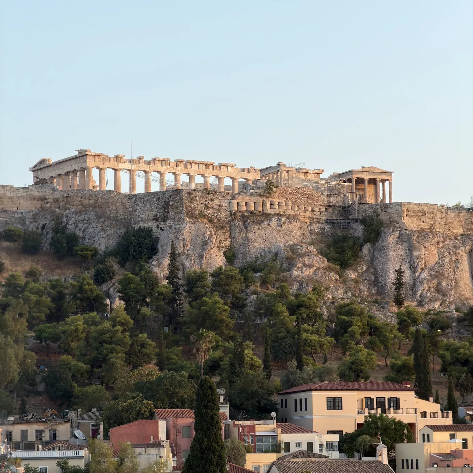 A scenic view of the Acropolis of Athens with the Parthenon atop a rocky hill, surrounded by green trees and historic buildings under a clear light blue sky.