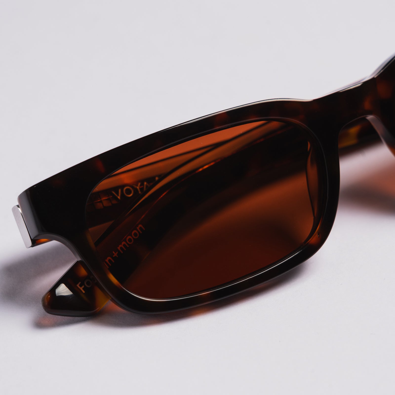 A close-up shot of a pair of sunglasses VOYA with a dark brown tortoiseshell frame and amber-brown lenses, resting on a light gray surface.