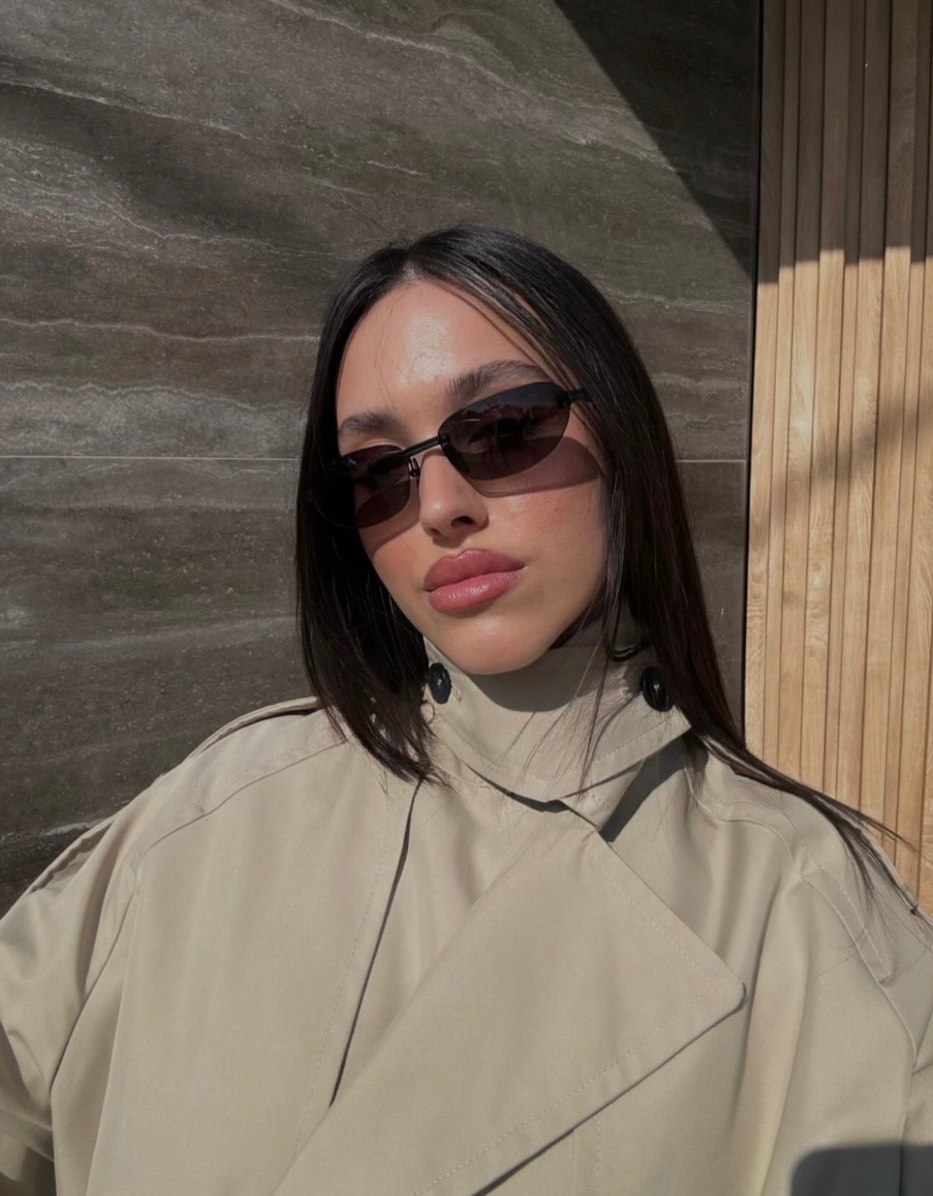 Woman wearing sunglasses and a beige coat against a textured wall.