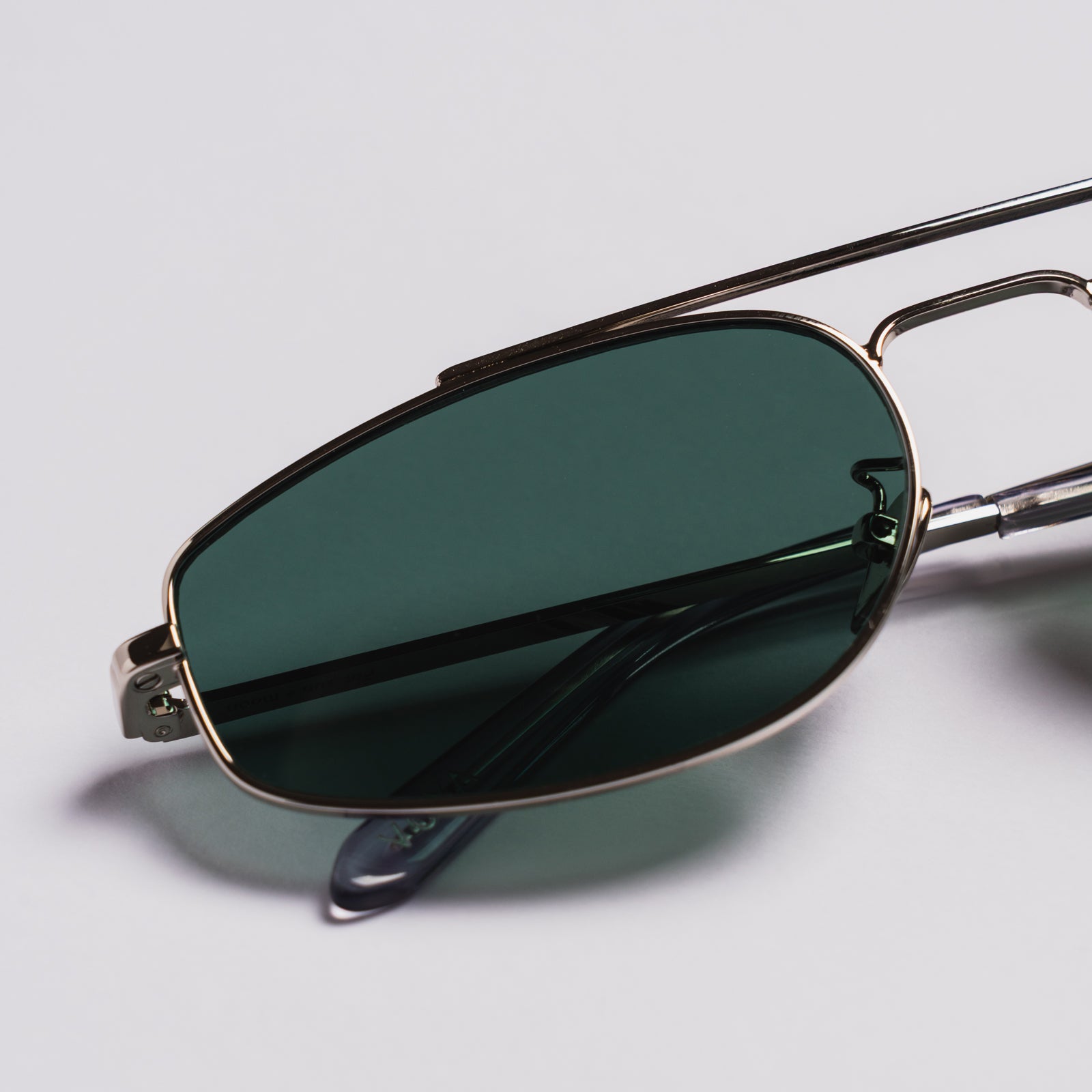Close-up of aviator-style sunglasses with dark green tinted lenses and a thin metallic frame, resting on a light gray background.