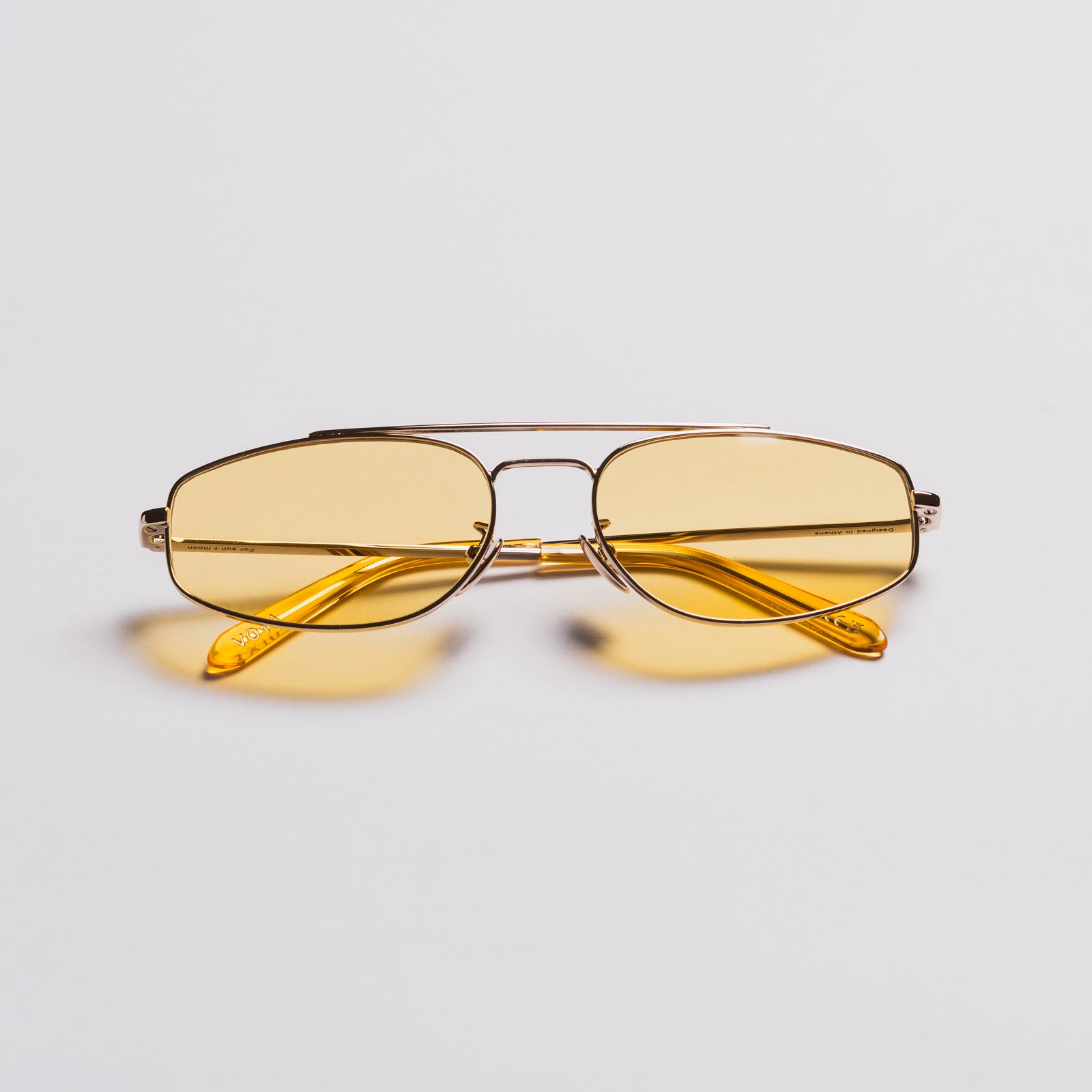 A pair of aviator-style sunglasses with yellow tinted lenses and a thin gold metal frame, resting on a light gray background with soft yellow reflections underneath.