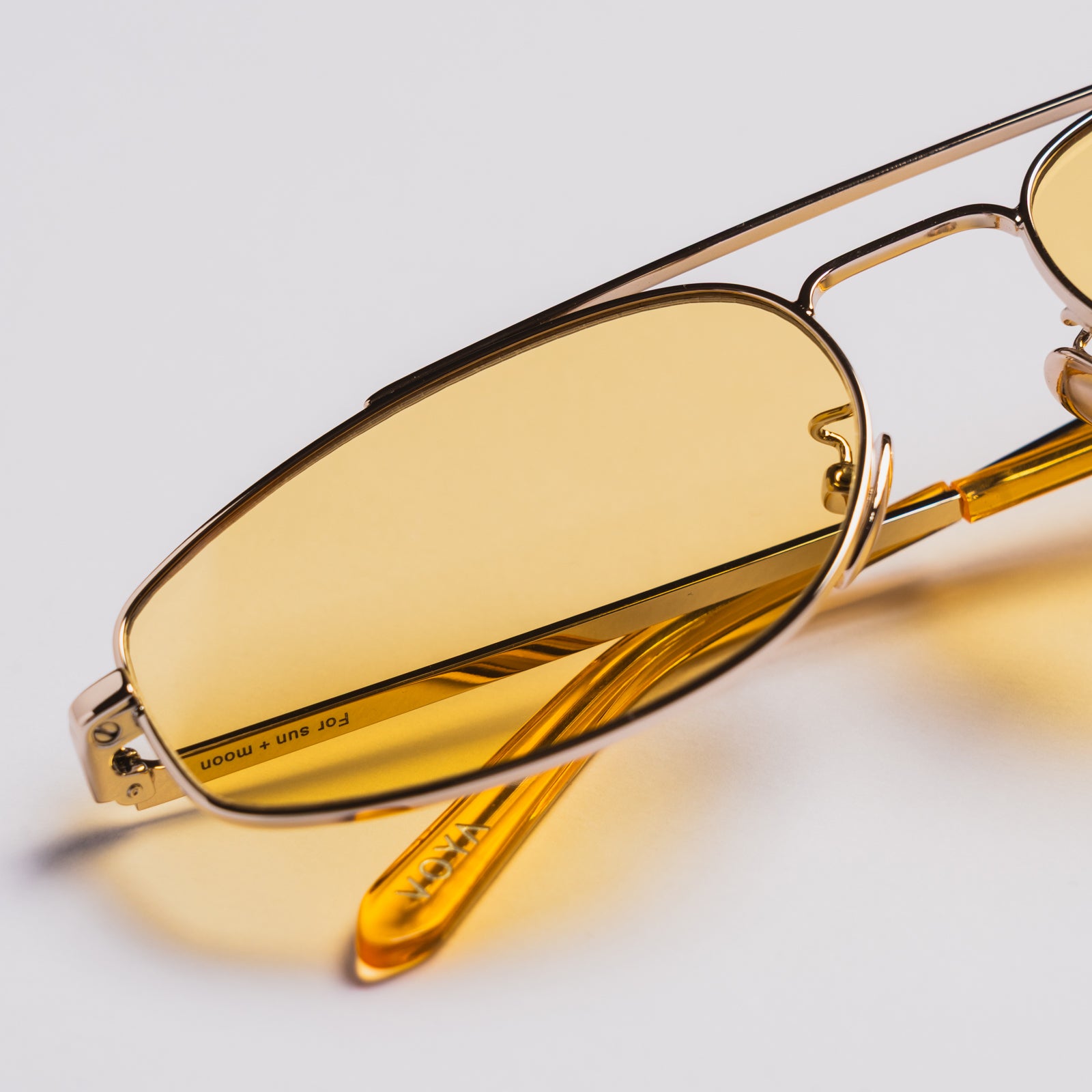 Close-up of aviator-style sunglasses with translucent yellow lenses and a thin gold metal frame on a light gray background.