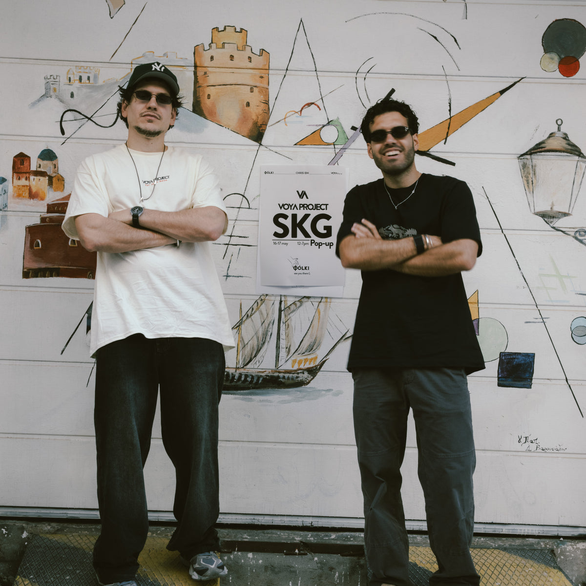 Two men standing in front of a white wall with illustrated drawings, holding a white sign that reads 'VON PROJECT SKG'.
