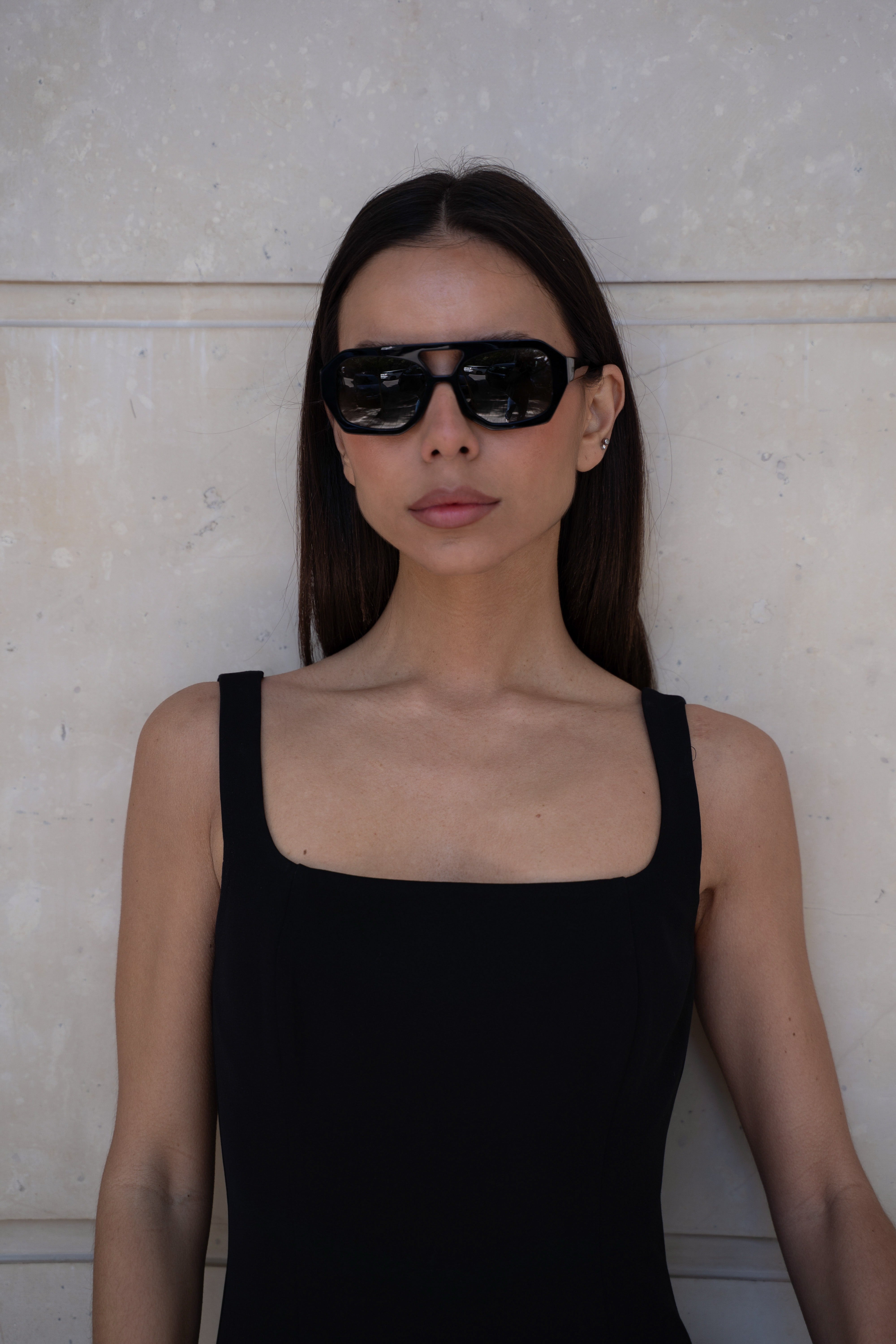 Woman wearing black sunglasses and a black sleeveless top against a light-colored wall.