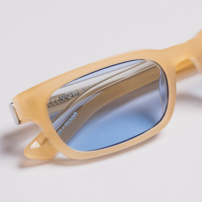 Close-up of a pair of sunglasses with a translucent pale yellow acetate frame and blue mirrored lenses, shown on a light gray background.