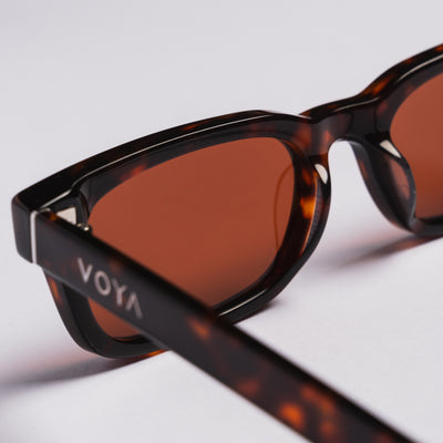 Close-up of VOYA sunglasses with a dark tortoiseshell acetate frame and warm brown lenses, resting on a light gray surface.