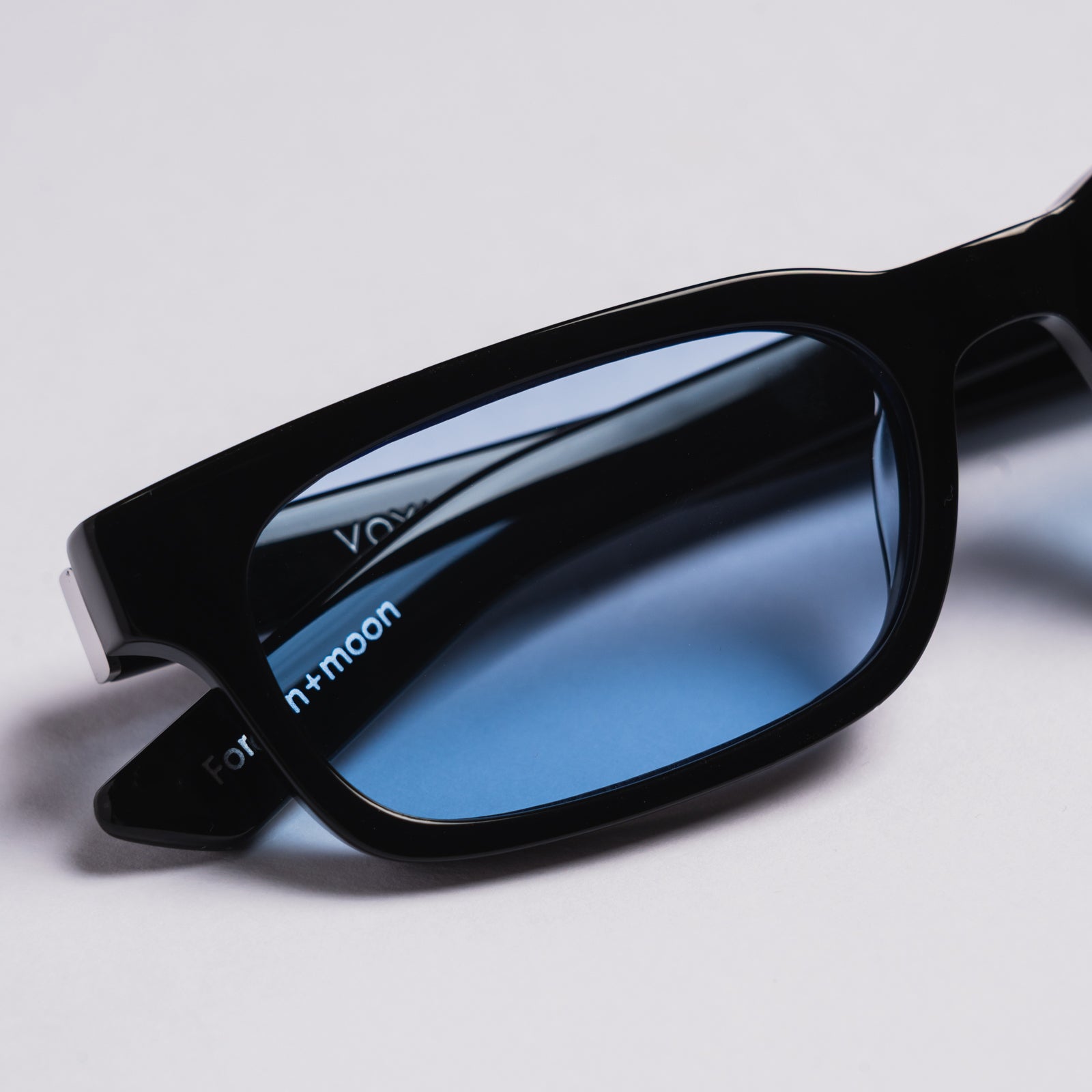 Close-up of a pair of sunglasses with a glossy black frame and blue-tinted lenses. The brand name 'n+moon' is printed in white on the lens, and part of 'Vony' is visible on the other side.