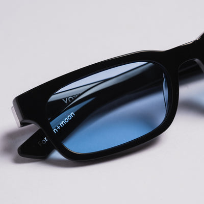 Close-up of a pair of sunglasses with a glossy black frame and blue-tinted lenses. The brand name 'n+moon' is printed in white on the lens, and part of 'Vony' is visible on the other side.
