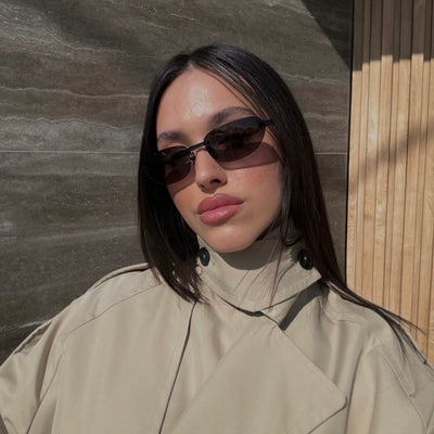 Woman wearing sunglasses and a beige coat against a textured wall.