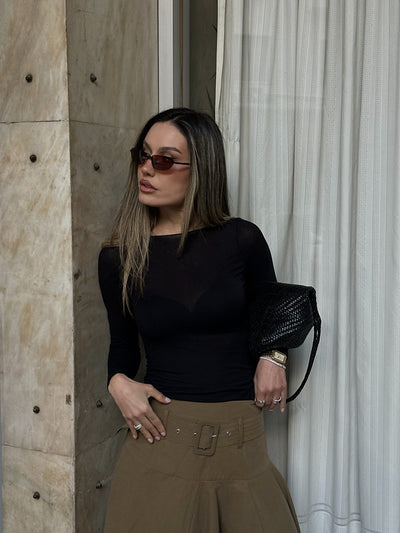 Woman in black top and brown pants standing against a textured wall with curtains.