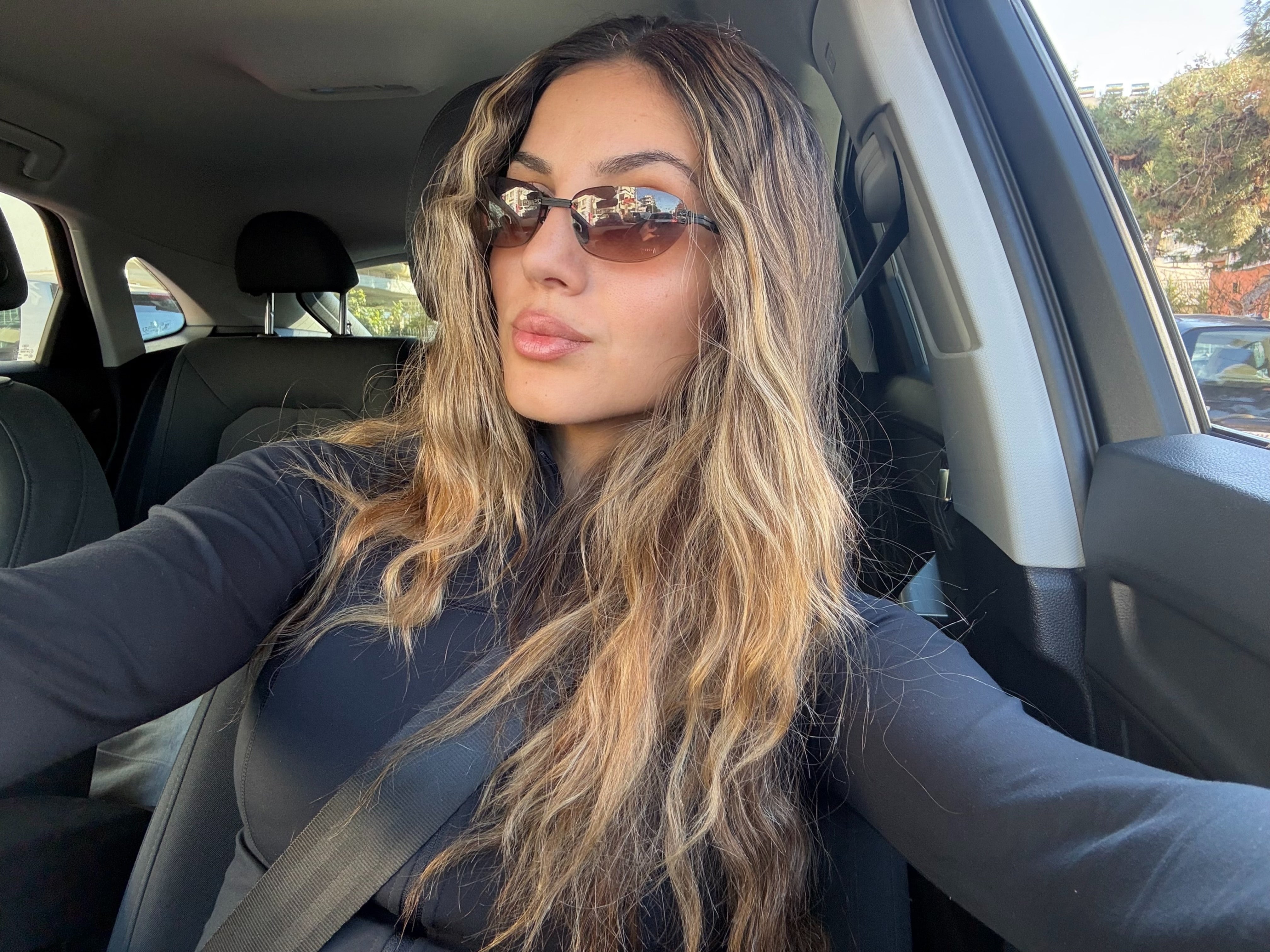 Woman sitting in a car wearing sunglasses and a dark jacket.