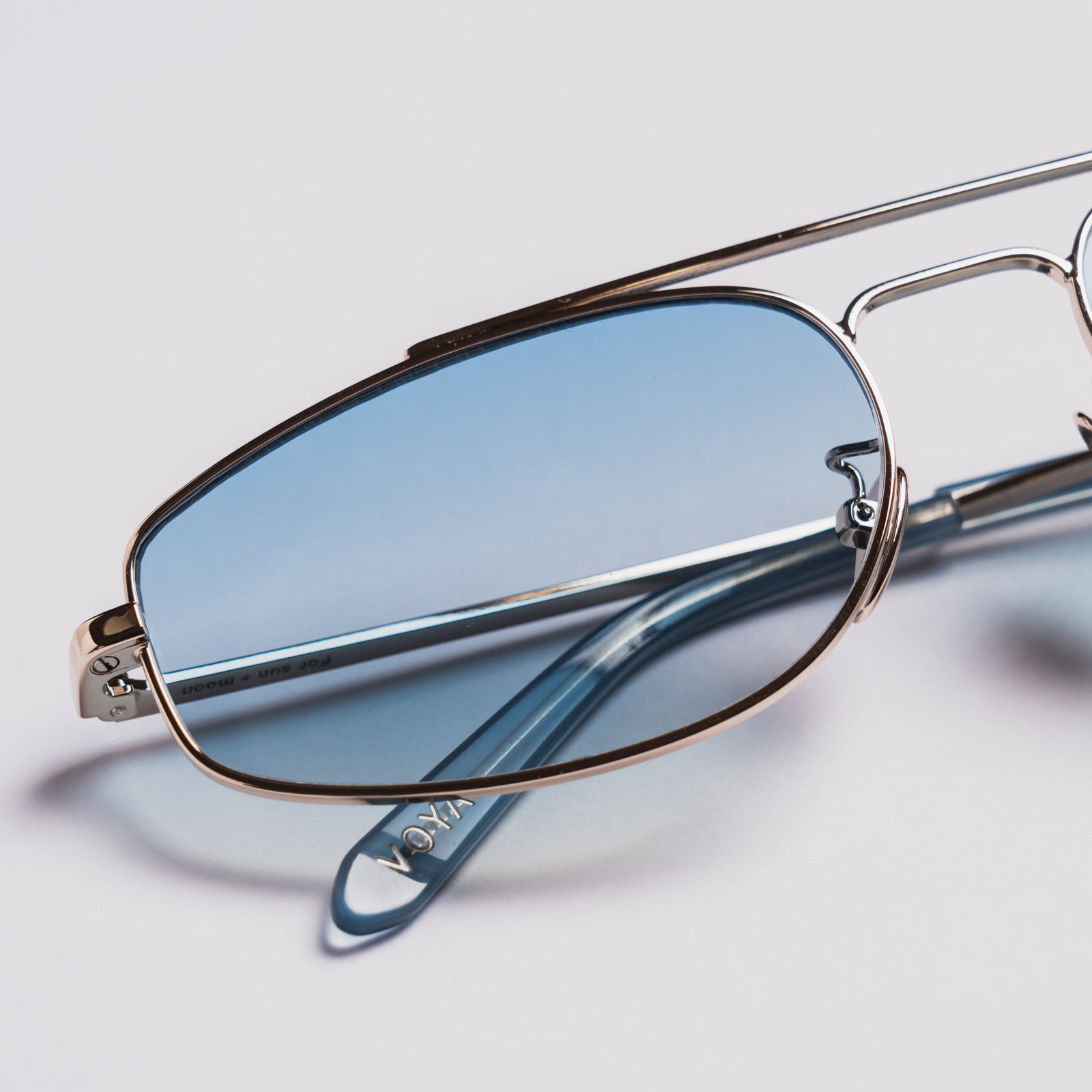 Close-up of a pair of eyeglasses with oval light blue lenses, a thin silver metal frame, and translucent blue temple arms on a light gray background.