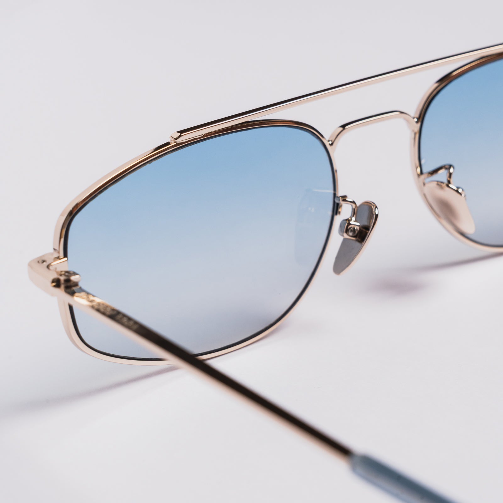 Close-up of aviator-style sunglasses with a thin gold-tone metal frame and light blue gradient lenses, shown on a clean white background.