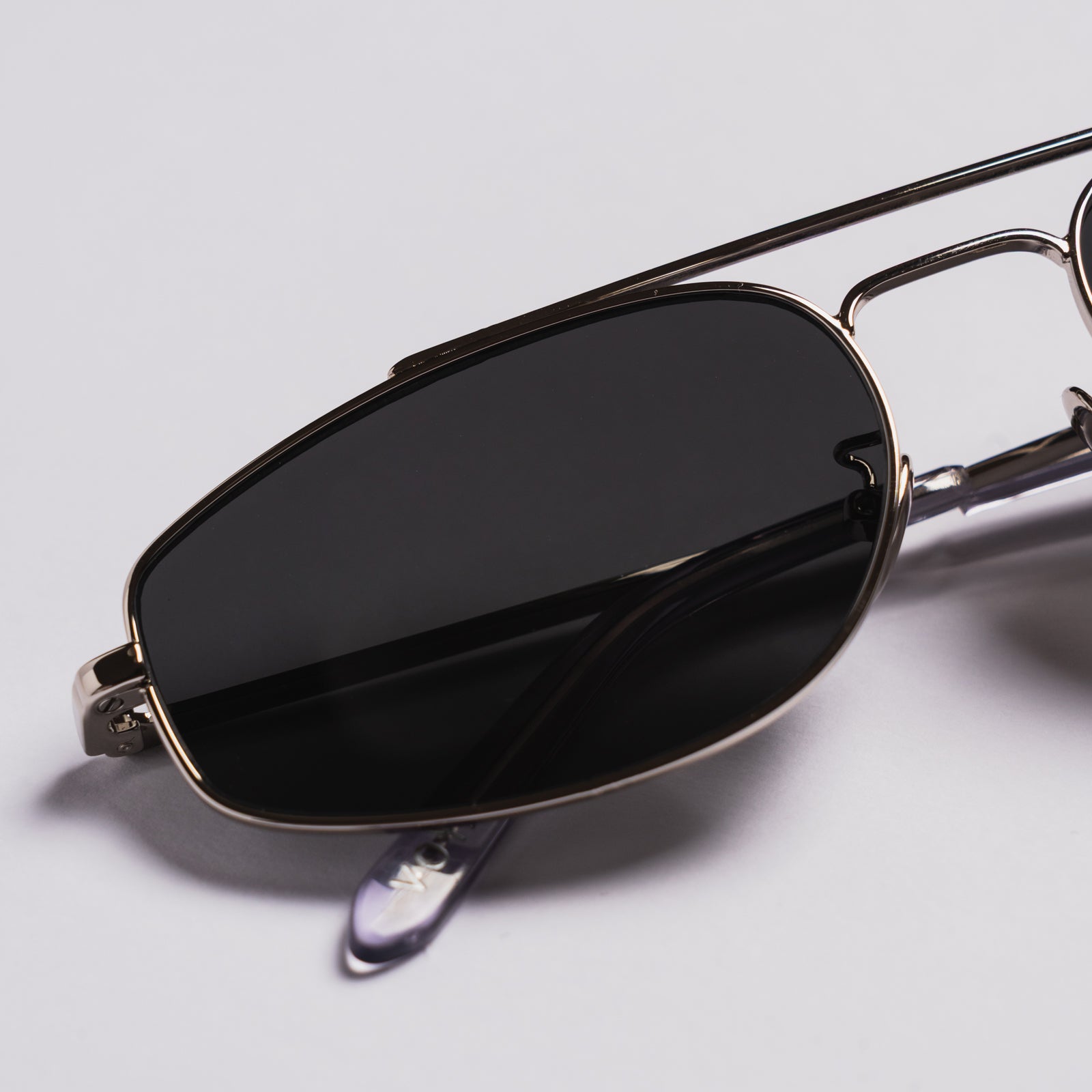 Close-up of a pair of aviator-style sunglasses with dark tinted lenses and a thin metallic frame, photographed on a light gray background.