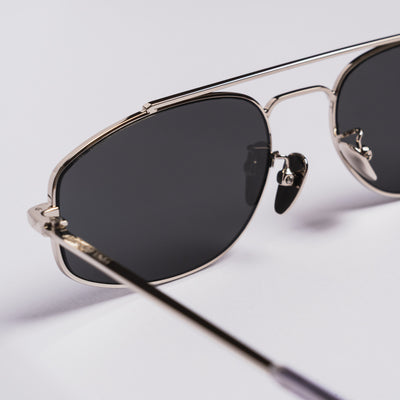 Close-up of aviator-style sunglasses with a thin silver metal frame and dark tinted lenses on a light gray background.