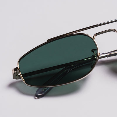 Close-up of aviator-style sunglasses with dark green tinted lenses and a thin metallic frame, resting on a light gray background.