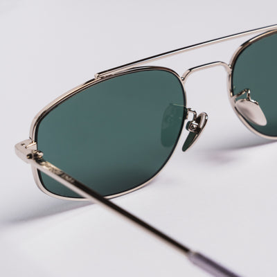 Close-up of aviator-style sunglasses with a thin silver metal frame and dark green tinted lenses on a light gray background.