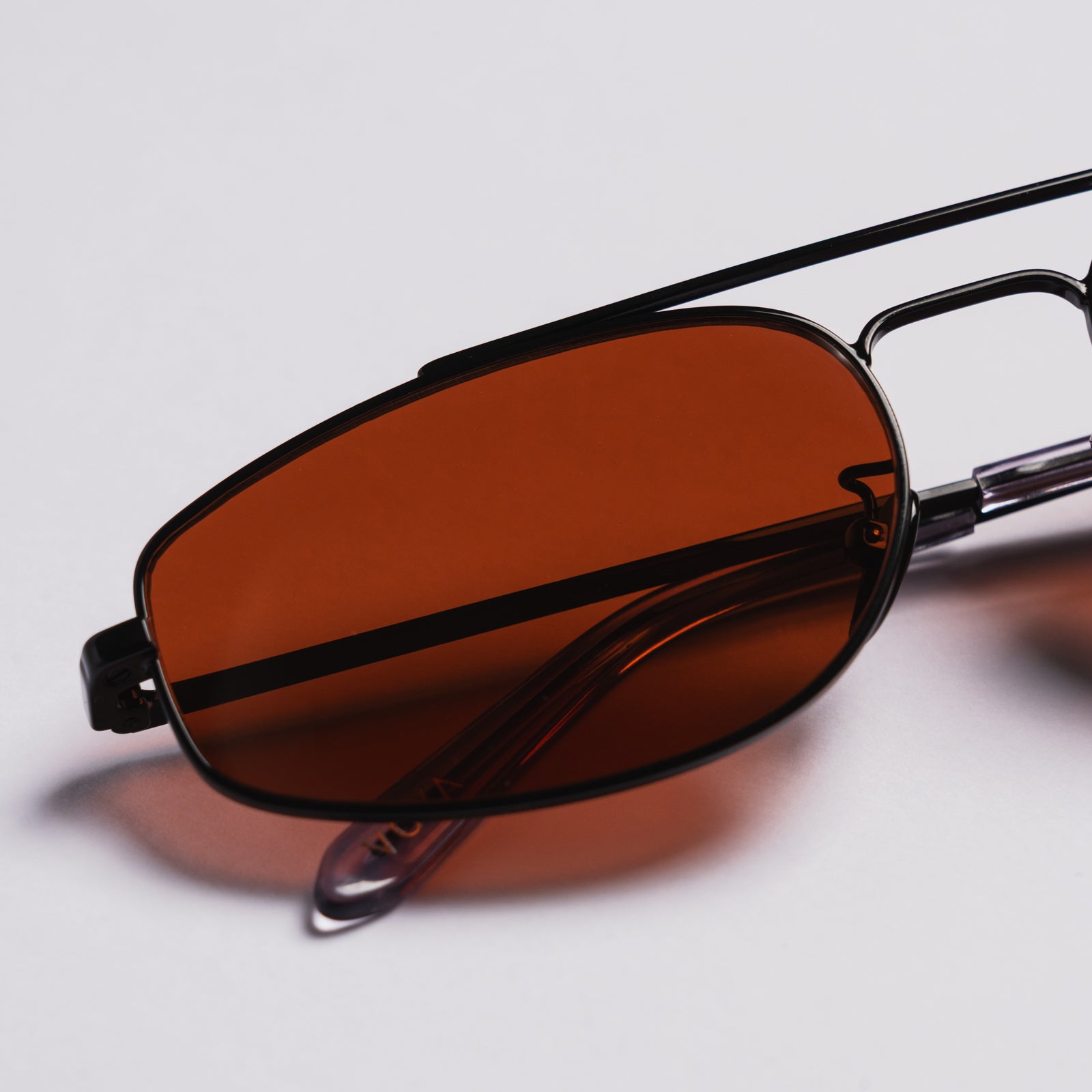 Close-up of a pair of sunglasses with a black metal frame and warm brown tinted lenses, resting on a light gray background.
