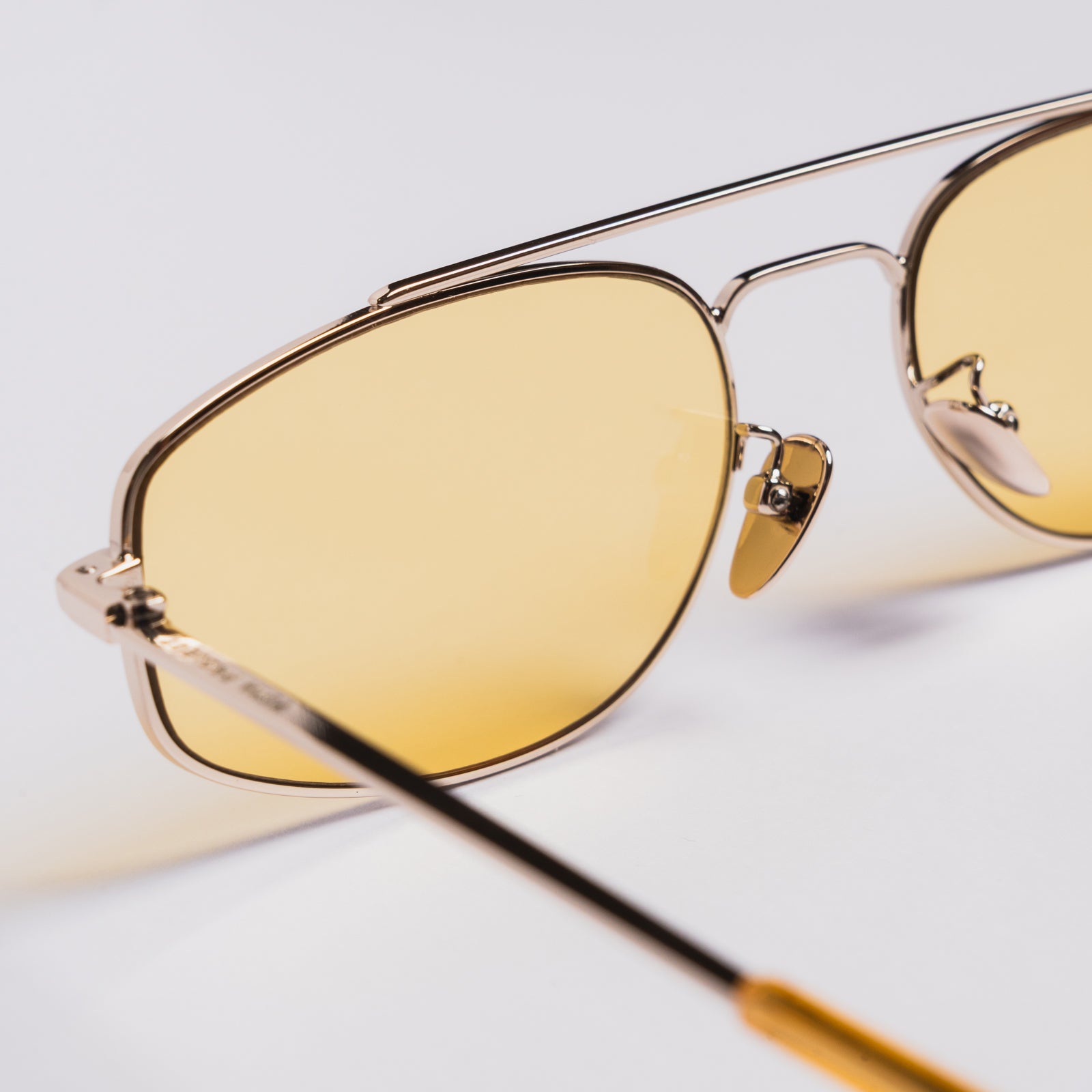 Close-up of aviator-style sunglasses with a thin gold-tone metal frame and warm yellow-tinted lenses, resting on a light neutral surface.