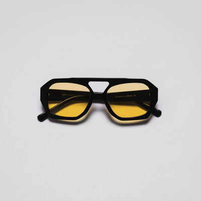A pair of sunglasses with black frames and yellow tinted lenses positioned against a white background.