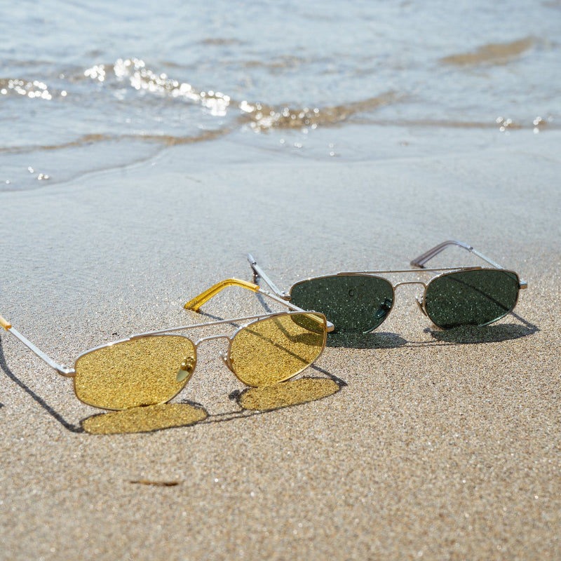 Two pairs of aviator sunglasses resting on wet sand near the shoreline, with ocean water in the background. The sunglasses have yellow, green, and dark gray lenses with gold-tone metal frames.